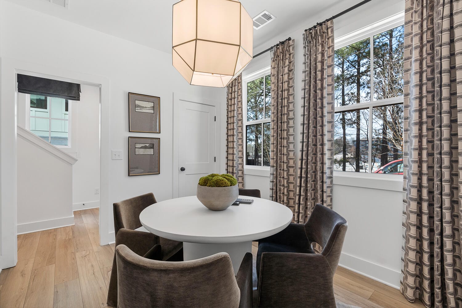 Small dining area with round white table, brown chairs, decorative bowl, wooden flooring, white walls, large windows with patterned curtains, geometric pendant light, open doorway with stairs, framed pictures.