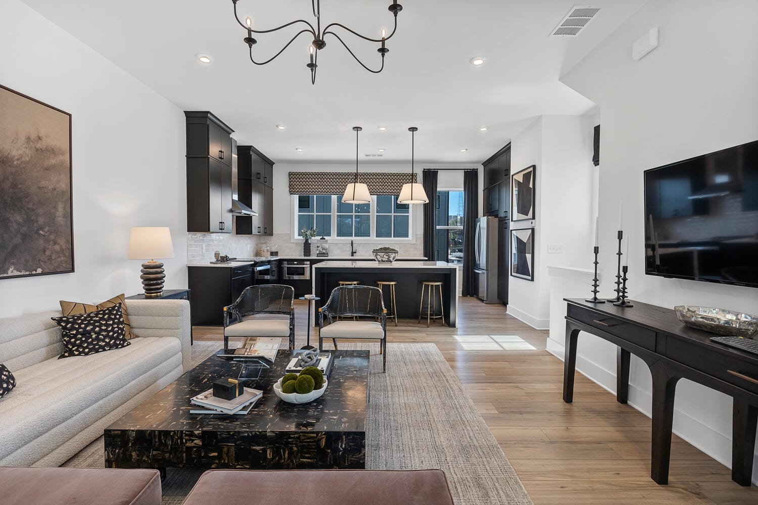 Open-concept living room and kitchen with light-colored sofa, patterned cushions, dark coffee table, chairs, large rug, dark cabinetry, stainless steel appliances, island with bar stools, chandelier, framed artwork, lamps, candlesticks, mounted TV.