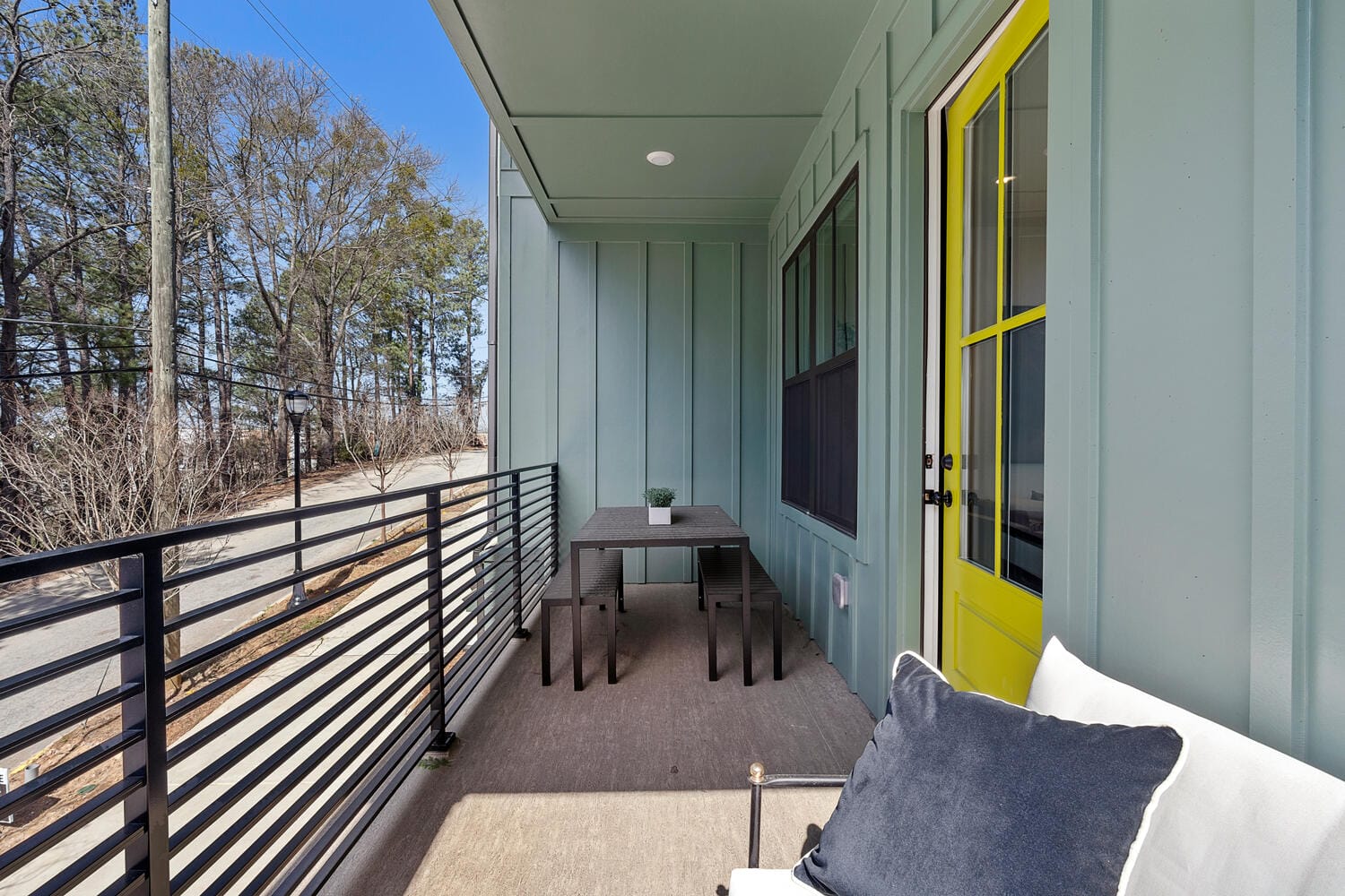 Outdoor balcony with table, benches, railing, yellow door, light blue-green walls, outdoor sofa with white cushions, trees, power lines.