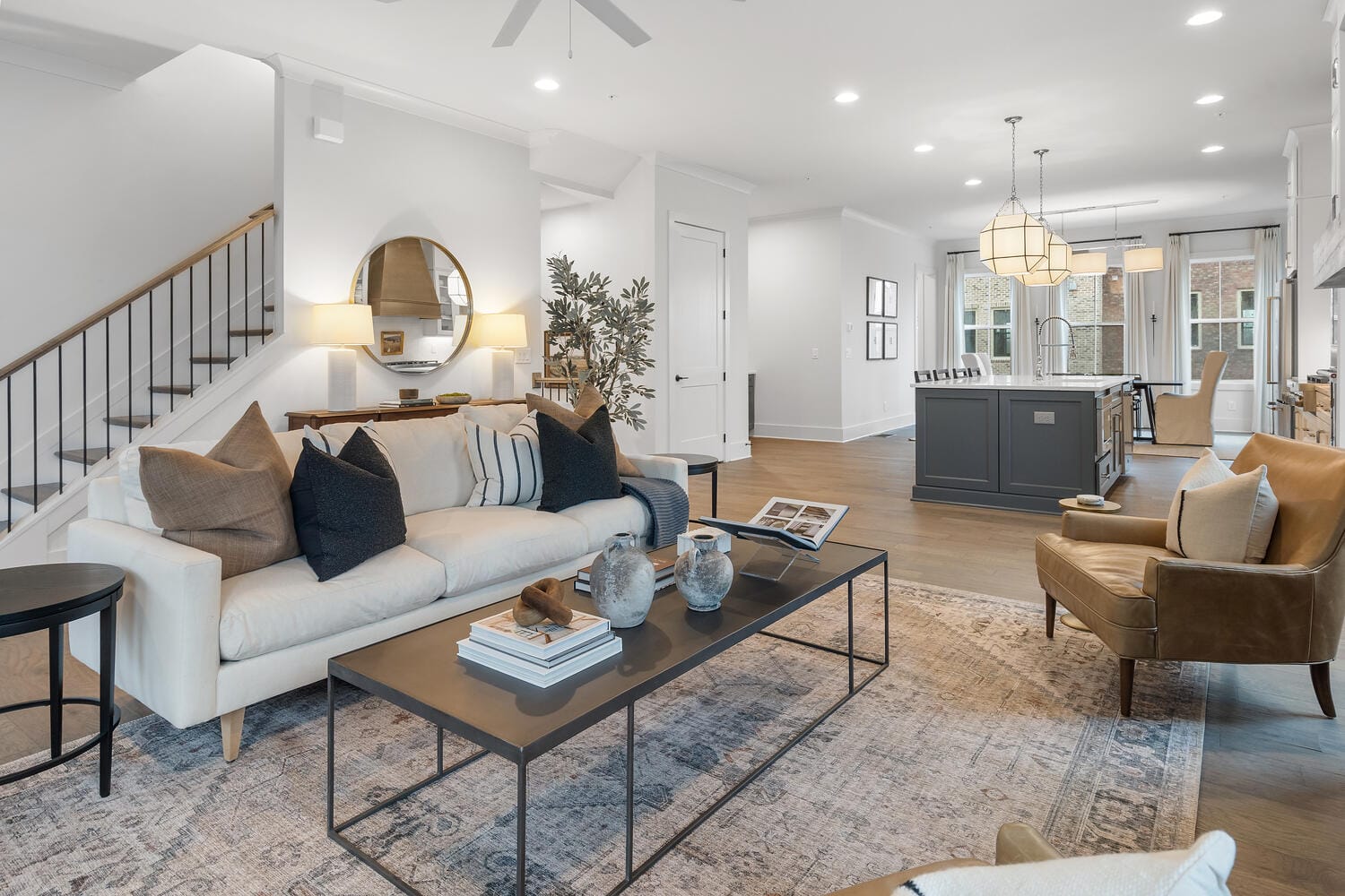 Modern open-concept living room and kitchen area with light-colored sofa, coffee table, armchair, staircase with railing, round mirror, wall-mounted lights, kitchen island with bar stools, pendant lighting, large windows, wooden flooring, and patterned rug.