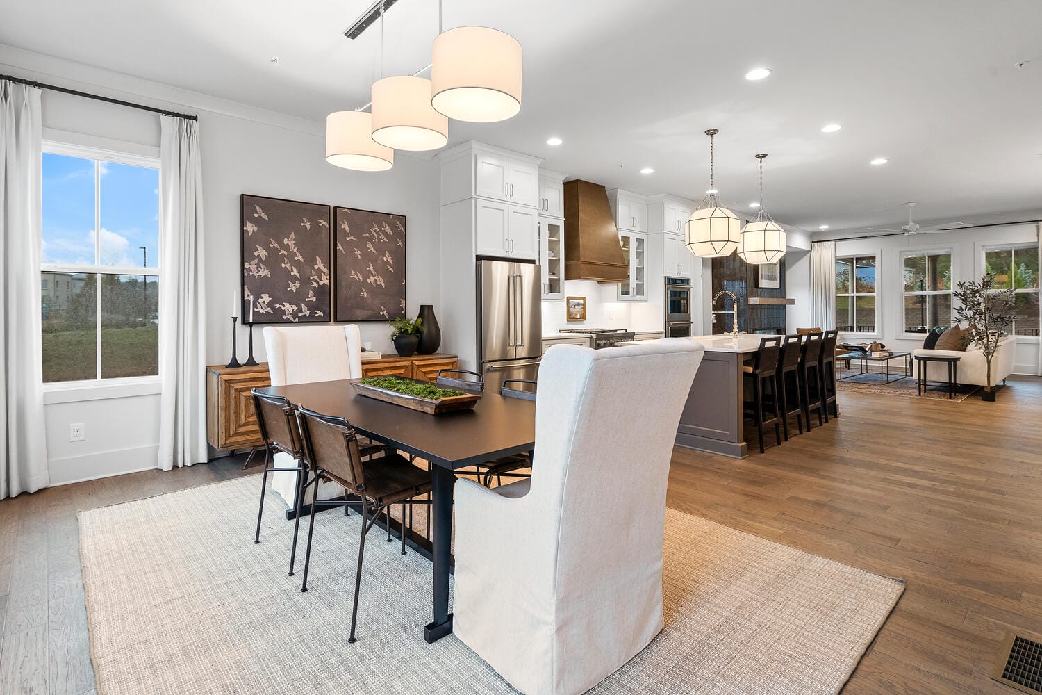 Open-concept kitchen and dining area, rectangular table with upholstered and wooden chairs, three pendant lights above table, sideboard with artwork, white cabinetry, stainless steel appliances, island with seating for six, large windows allowing natural light.