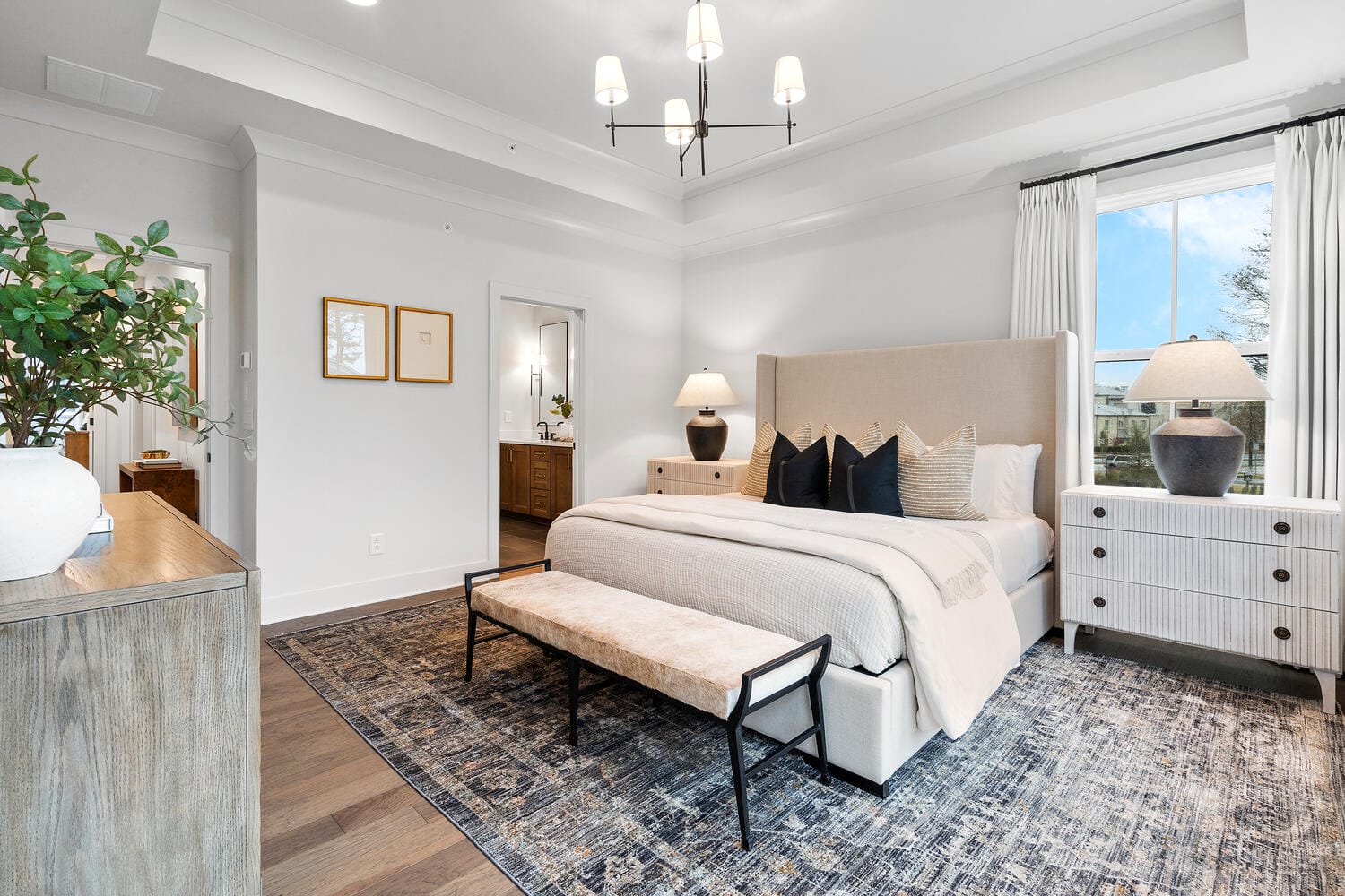 Modern bedroom with large beige upholstered bed, multiple pillows, bench at foot of bed, nightstands with lamps, dresser with decorative items, window with white curtains, patterned rug, white walls, framed pictures, and open doorway.