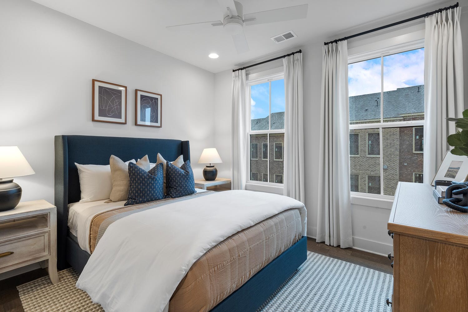 Bedroom with a large bed in the center, dark blue headboard, white and beige bedding, decorative pillows, two bedside tables with lamps, large windows with white curtains, white walls, three framed pictures above the bed, ceiling fan, small plant on bedside table, electronic device or clock on dresser.