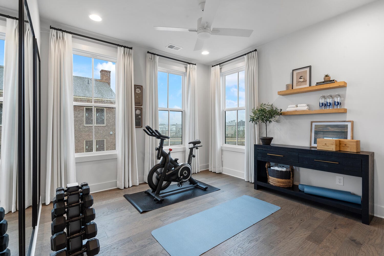 Home gym with exercise bike, dumbbells, yoga mat, console table, and large windows.