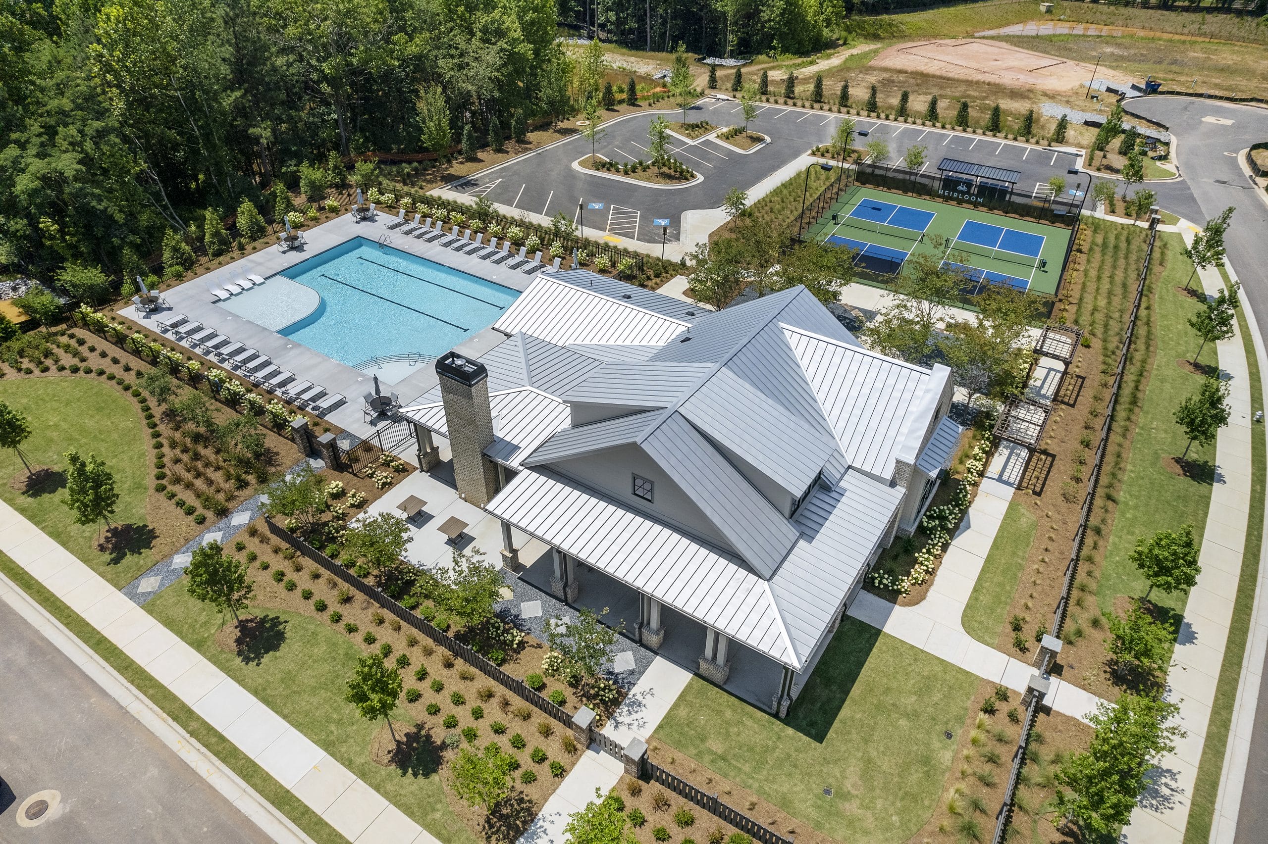 Aerial view of community recreational area with large swimming pool, clubhouse with metal roof, multiple tennis courts, landscaped gardens, and parking spaces, showcasing amenities.