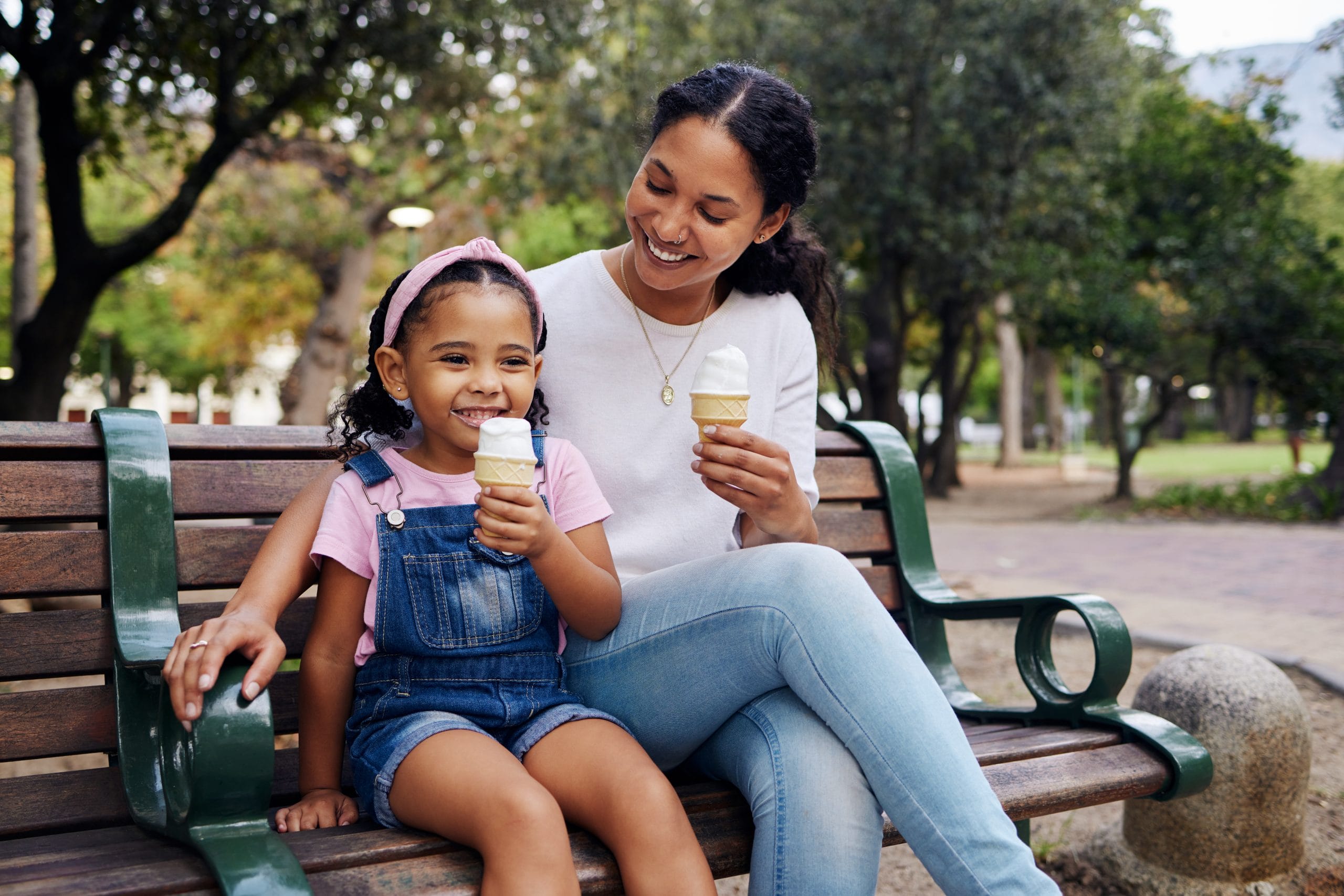 Black family, park and ice cream with a mother and daughter bonding together while sitting on a bench outdoor in nature. Summer, children and garden with a woman and girl enjoying a sweet snack.