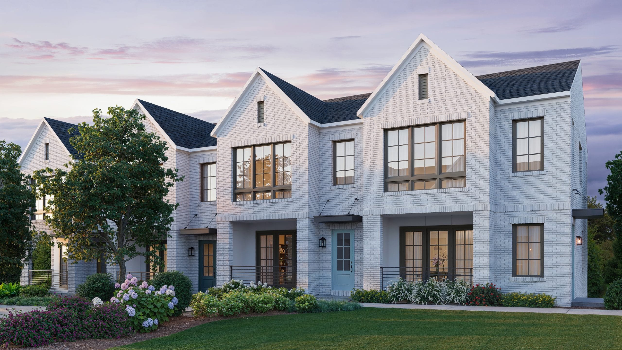 Modern two-story townhouse with white brick exterior, large windows, gabled roofs, well-maintained lawn, shrubs, flowering plants, and trees, during sunset or sunrise.