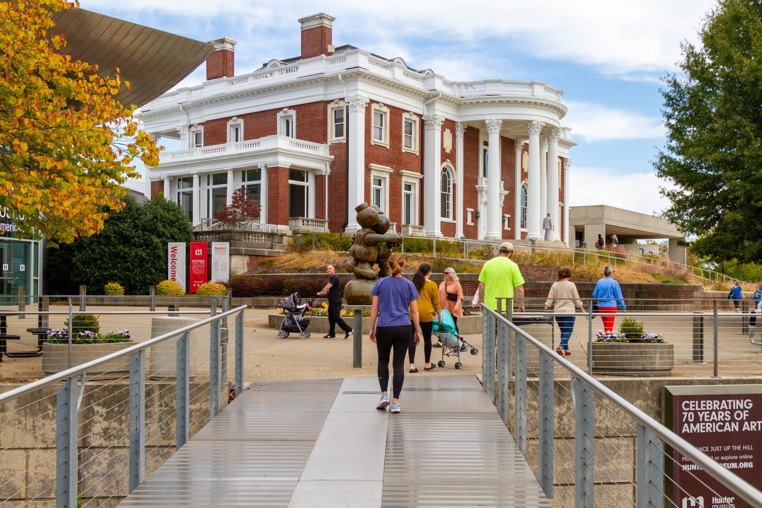 Group of people walking on a bridge towards a large historic building with white columns and red brick walls. Fountain with stacked stones, autumn foliage, and clear skies.