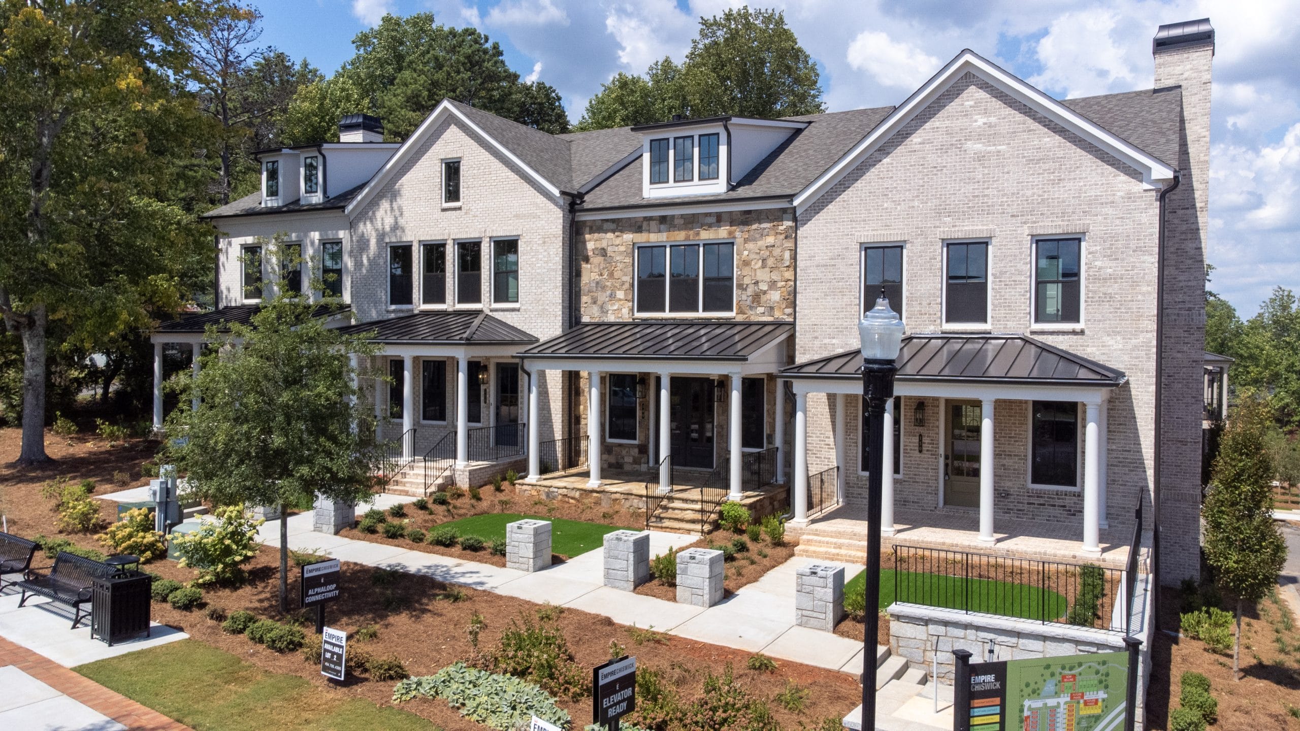 Multi-story townhouses with well-maintained landscaping, brick and stone exteriors, large windows, covered porches, small lawns and shrubs, trees, sidewalk, and streetlamp.