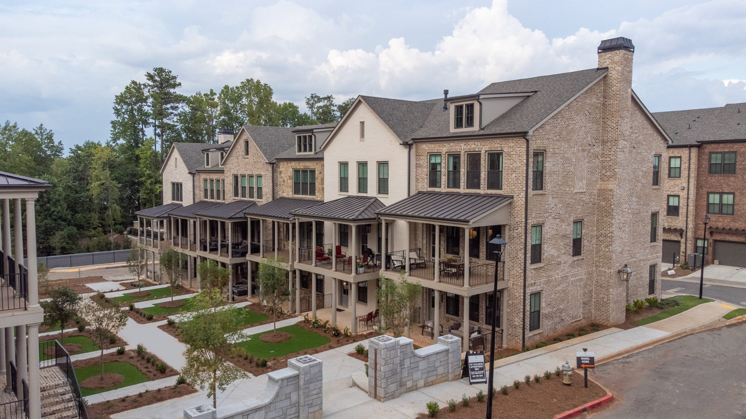 Residential building with brick exterior and multiple balconies, modern architectural design with large windows, well-maintained landscape with small trees and shrubs, neatly arranged pathways and retaining walls, part of a residential complex or neighborhood with another similar building visible in the background.