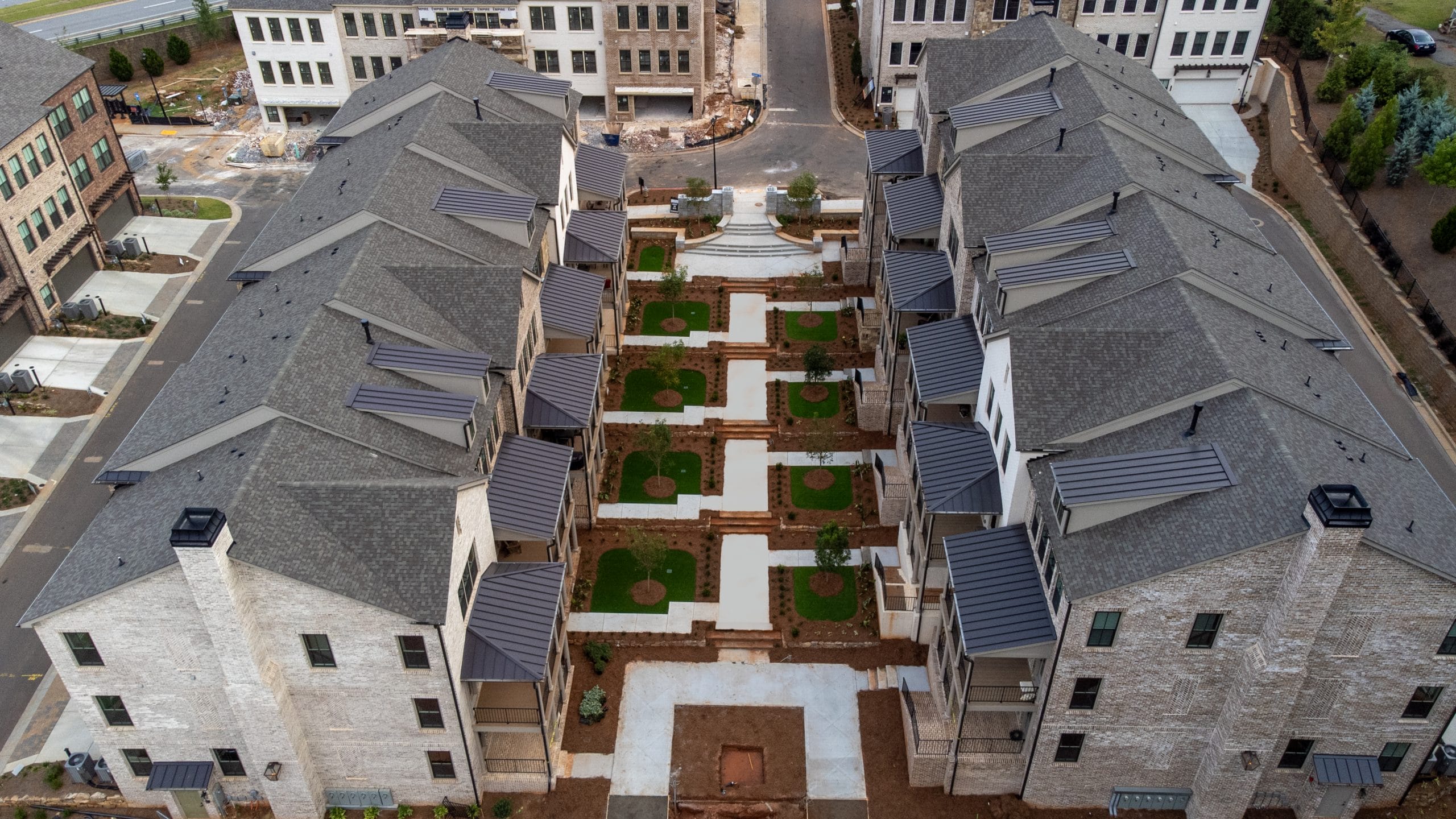 Aerial view of a residential complex with two rows of multi-story buildings facing each other, gray roofs and light-colored exteriors, landscaped area with pathways, green lawns, and small trees or shrubs arranged symmetrically, newly constructed or under construction with exposed soil and construction materials visible in the background.