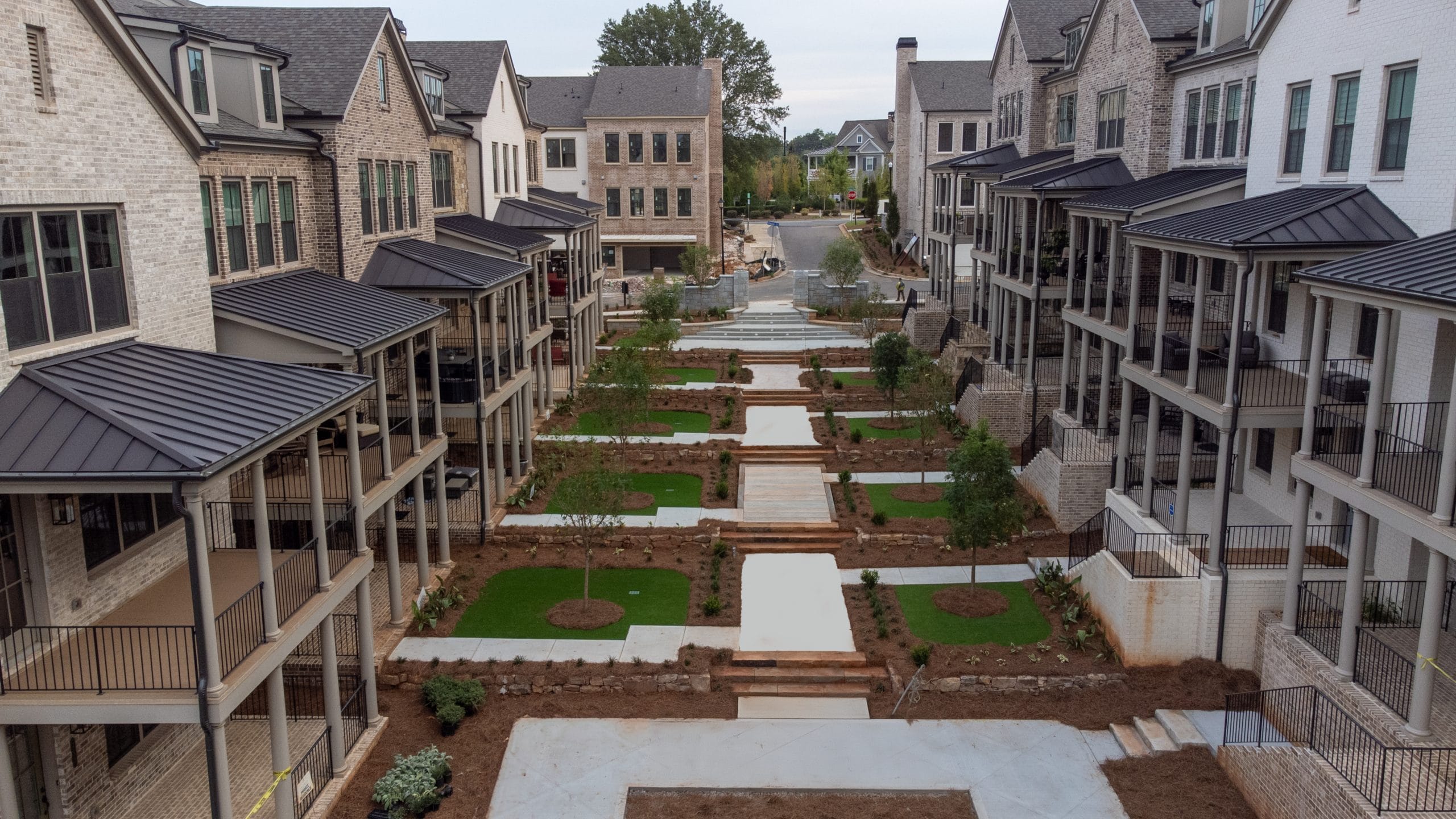 Residential area with two rows of multi-story townhouses facing each other, brick exteriors and large windows, some featuring balconies or porches, landscaped courtyard with pathways, stairs, and greenery.