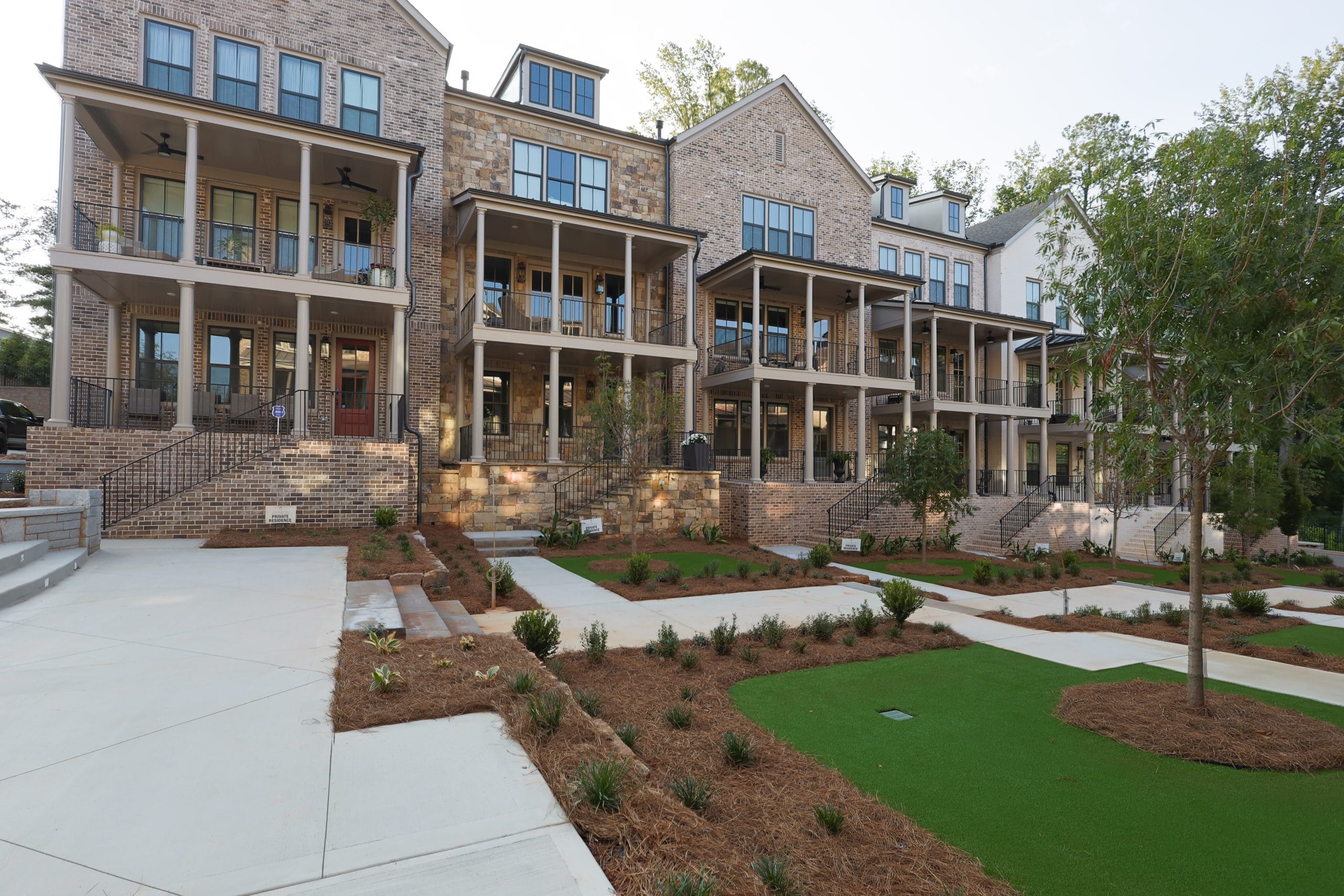 Row of multi-story townhouses with brick exteriors and large balconies, landscaped area with neatly trimmed bushes, small trees, patches of grass, paved walkways, and steps leading up to the entrances.