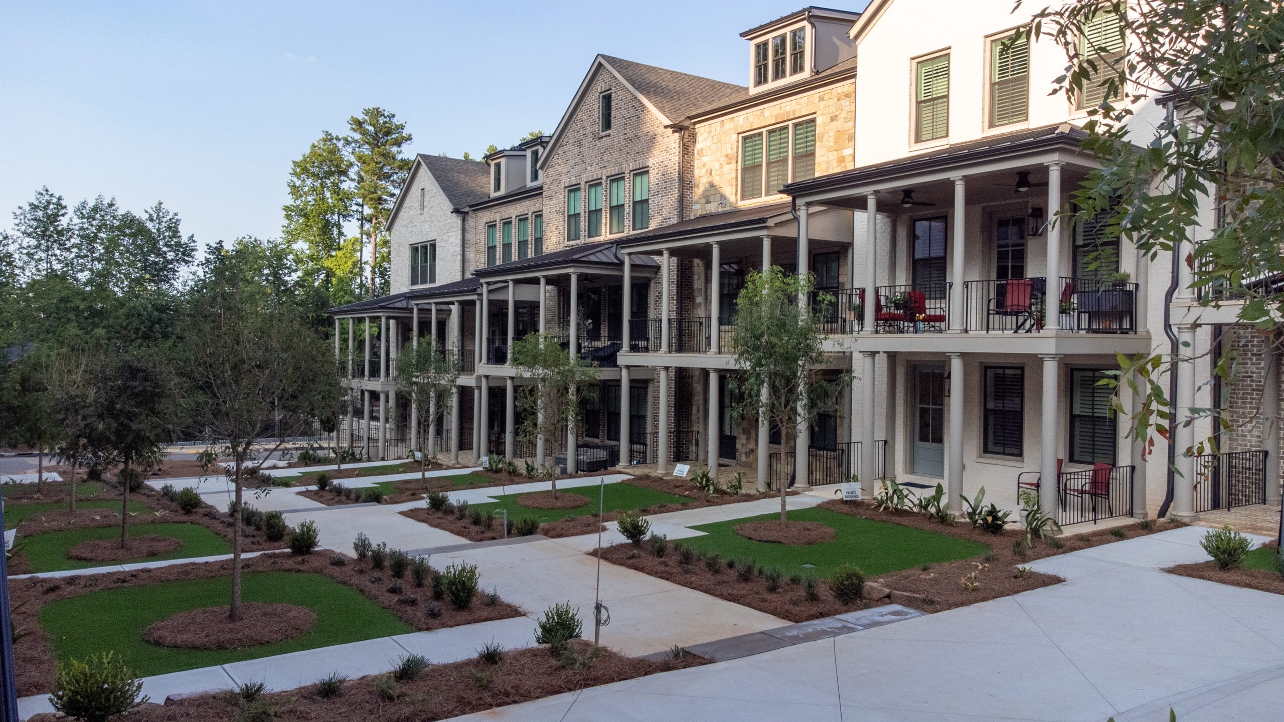 Multi-story residential buildings with balconies and porches, surrounded by landscaped areas with grass, trees, and shrubs, paved walkways and driveways in front of the buildings.