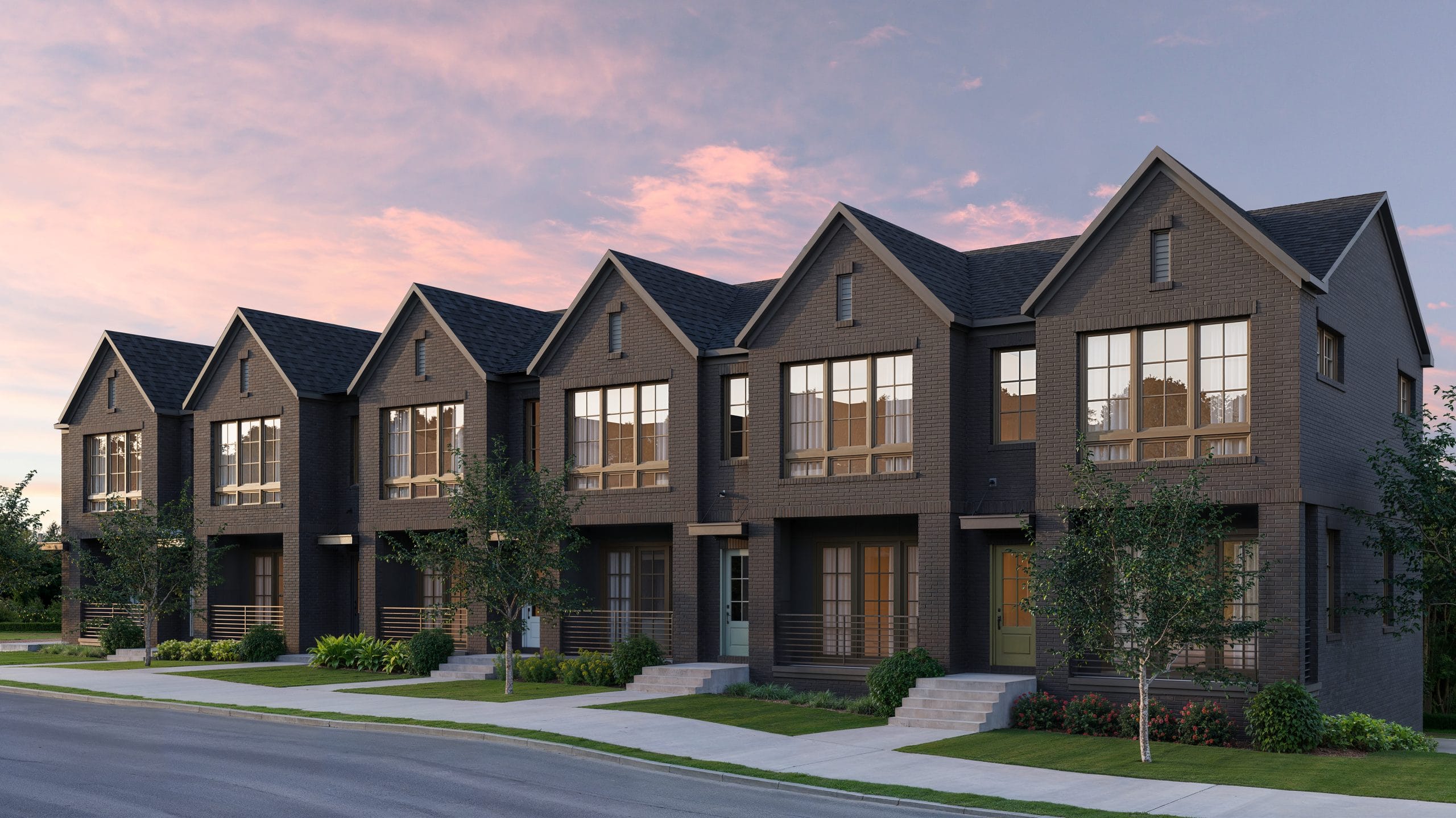Row of modern townhouses with dark brick exteriors, large windows, small front porches, neatly maintained landscaping with trees and shrubs, partly cloudy sky with pinkish hue, clean and well-maintained street.