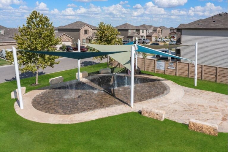 A small splash pad with water jets, covered by shade sails, sits on a stone surface in a grassy park area. Nearby are benches, trees, and surrounding houses under a blue sky with scattered clouds.